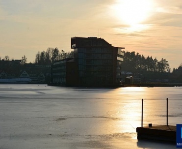 Zach&oacute;d słońca nad Mikołajkami - Zimowy zach&oacute;d słońca nad zamarzniętym jeziorem w Mikołajkach, z widokiem na hotel w tle, typowe dla Mazur.